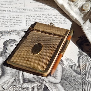 May include: A rectangular gold-tone metal notebook with a textured cross-hatch finish and a smooth oval center plate. A small pencil with a wooden body and a gold-tone metal tip is held in a side loop attached to the spine of the notebook.