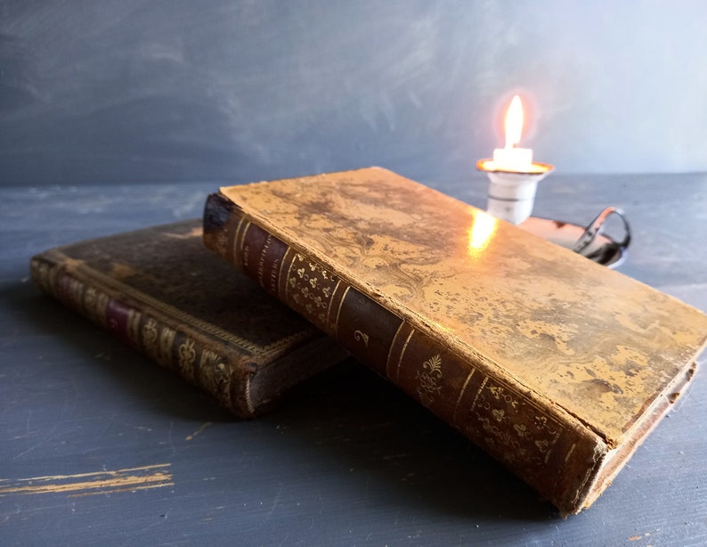 Op de afbeelding: Twee antieke boeken met goud bedrukte omslagen en versleten leren ruggen. Op de achtergrond staat een brandende kaars op een tafel.