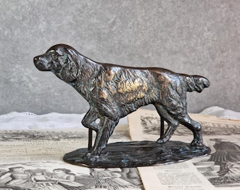 Op de afbeelding: Een bronskleurig metalen beeldje van een jachthond in staande positie. Het figuur heeft een getextureerde, verweerde afwerking met accenten van lichtere goudtinten op de vacht, geplaatst op een ovale voet die wordt ondersteund door twee staven.