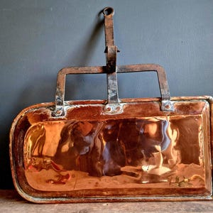 May include: Antique copper pan with a rectangular shape and rounded corners. It features a riveted metal handle and a hanging loop. The copper surface has a reflective, polished finish, showing signs of age.