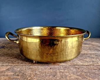 Op de afbeelding: Een gehamerde messing plantenbak met twee handvatten en drie poten. De plantenbak is rond en heeft een glanzende gouden afwerking.