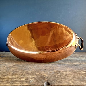 May include: A copper-coloured, oval-shaped bowl with two small handles. The bowl has a reflective surface and sits on a weathered wooden surface. The background is a solid, dark blue colour. This decorative bowl is suitable for serving.