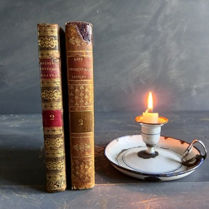 Op de afbeelding: Twee antieke boeken met gouden en rode accenten en de tekst "ALLUSIONS LITTERAIRES DE LA PICARDIE" en "LORD CHESTERFIELD'S LETTERS" op de ruggen. De boeken staan op een houten oppervlak naast een witte kaarsenstandaard met een brandende kaars.