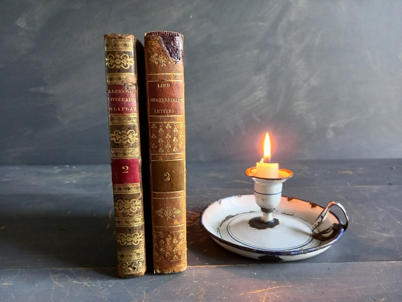 Op de afbeelding: Twee antieke boeken met gouden en rode accenten en de tekst "ALLUSIONS LITTERAIRES DE LA PICARDIE" en "LORD CHESTERFIELD'S LETTERS" op de ruggen. De boeken staan op een houten oppervlak naast een witte kaarsenstandaard met een brandende kaars.