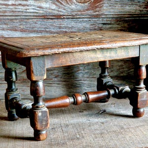 Peut inclure: Un petit tabouret en bois antique avec un dessus rectangulaire et quatre pieds tournés. Le bois a un aspect vieilli et patiné avec des veines visibles et des variations de couleur, allant du brun clair au brun foncé.