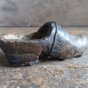 May include: A dark brown wooden clog with a worn and weathered surface. The clog is missing a piece on the side, revealing the hollow interior.