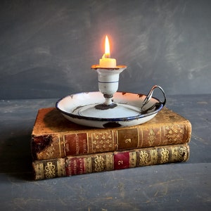 Op de afbeelding: Een witte emaille kaarsenstandaard met een blauwe rand staat op twee antieke boeken. De kaarsenstandaard heeft een handvat en een kleine witte kaars brandt erin.