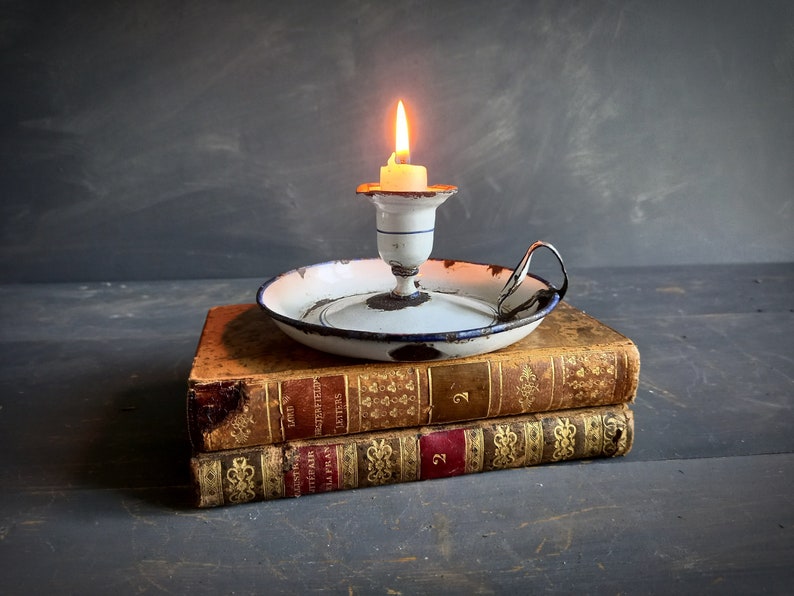 Op de afbeelding: Een witte emaille kaarsenstandaard met een blauwe rand staat op twee antieke boeken. De kaarsenstandaard heeft een handvat en een kleine witte kaars brandt erin.