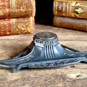 Peut inclure: Un encrier vintage argenté avec un design orné et un dessus rond de couleur cuivre. L'encrier est posé sur une surface en bois.