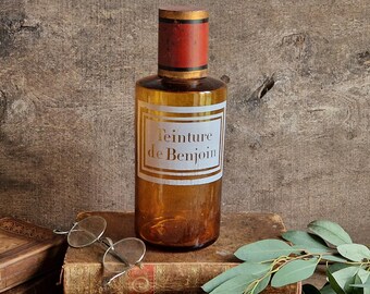 May include: An amber glass apothecary bottle with a red and gold cylindrical stopper. The bottle features a white rectangular label with a gold border and the black text Teinture de Benjoin printed in the centre of the front surface.