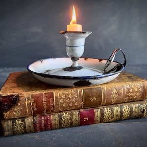 Op de afbeelding: Een witte en blauwe emaille kaarsenstandaard met een handvat staat op twee antieke boeken. De kaarsenstandaard heeft een versleten, rustieke uitstraling en de boeken zijn gebonden in leer met gouden letters. De kaars is aangestoken en geeft een warm licht.