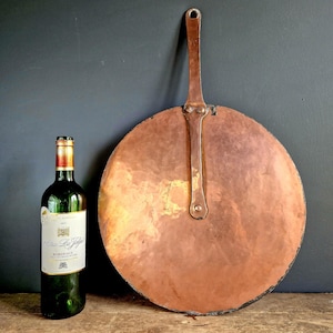 Op de afbeelding: Een grote, ronde koperen pan met een lange handgreep, naast een groene wijnfles. De pan heeft een gehamerde textuur en een donkere patina. De wijnfles heeft een wit etiket met gouden accenten. De achtergrond is donkergrijs.