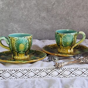 May include: Two ceramic cups and matching saucers featuring a raised leaf pattern in shades of green and yellow, with a light blue interior. A pair of ornate silver-toned metal tongs with intricate embossed detailing rests beside the cups.