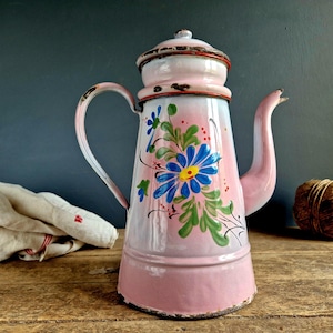 以下が含まれることがあります： 花柄のヴィンテージピンクエナメルコーヒーポット。ポットは、黄色い中心、緑の葉、赤いアクセントが付いた大きな青い花が特徴です。白いハンドルと注ぎ口、お揃いの蓋が付いています。