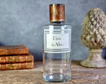 May include: A clear glass apothecary bottle with a cylindrical shape and a metallic, distressed copper-coloured cap. The front features a rectangular white label with a double-line border and the black printed text Tinc: de Aloe.