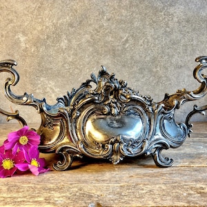 Peut inclure: Un centre de table en métal argenté orné de volutes complexes, de poignées incurvées et d'un motif central en relief. Le récipient repose sur quatre pieds décoratifs et est accompagné d'un bouquet de fleurs cosmos rose vif.