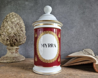 May include: A decorative apothecary jar with a white lid and base, and a red body. The jar is adorned with gold accents and the word "MYRRA" in a white oval. A stone sculpture and an open book with glasses are in the background.