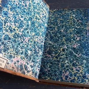 Op de afbeelding: Close-up van de marmeren omslag van een vintage boek. De omslag is diepblauw met swirls van geel en roze. De tekst op de rug staat "REG. PAROCH. S. SULPITII."