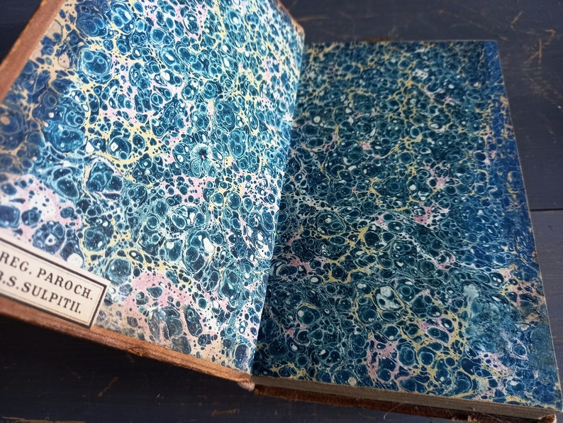 Op de afbeelding: Close-up van de marmeren omslag van een vintage boek. De omslag is diepblauw met swirls van geel en roze. De tekst op de rug staat "REG. PAROCH. S. SULPITII."