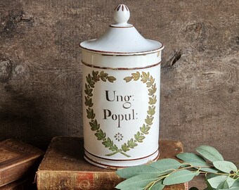 May include: A white cylindrical metal canister with a removable lid featuring a brown rim and a finial. The front displays a green painted laurel wreath surrounding the black text "Ung: Popul:" with a small decorative star symbol below.