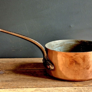 May include: A vintage copper saucepan with a long handle. The saucepan has a shiny, burnished finish and is sitting on a wooden surface.