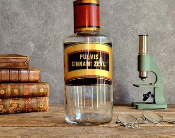 May include: A clear glass apothecary bottle with a red and gold lid, featuring a black and gold label that reads PULVIS CINNAM: ZEYL:. The bottle is cylindrical with a wide base and a narrow neck, displaying a vintage aesthetic with visible wear.