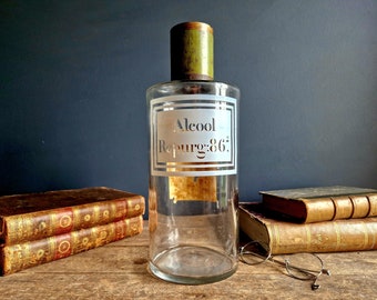 May include: A clear glass bottle with a green stopper and a white label that reads "Alcool Repurg: 86°". The bottle is sitting on a wooden surface with some old books and a pair of spectacles.