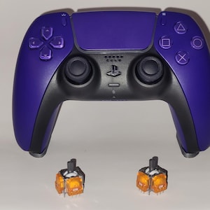 May include: A purple and black wireless video game controller featuring a central touchpad, two black analog sticks, a directional pad, and four action buttons, accompanied by two small orange replacement analog stick modules.
