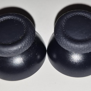 May include: Two black replacement analog thumbsticks for a video game controller. Each thumbstick features a smooth, rounded top surface with a textured, cross-hatched grip pattern around the outer edge and a solid, flared base for attachment.
