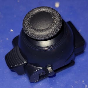 May include: A black joystick thumbstick with a textured, circular top. The base is a matte black with a rounded shape and angular sides. The thumbstick is designed for gaming controllers.