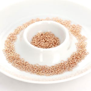 May include: A white ceramic dish filled with small, rose gold jump rings. The jump rings are arranged in a circular pattern around the edge of the dish.