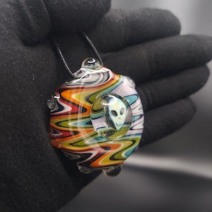 May include: A colorful glass pendant with a swirling pattern of red, orange, yellow, green, blue, black, and white. The pendant features a clear glass circle with a gray alien face inside.