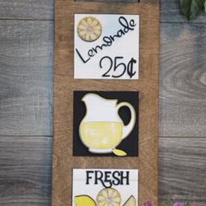 May include: Wooden lemonade stand sign with three panels. The top panel reads "Lemonade 25¢" with a lemon illustration. The middle panel shows a yellow pitcher. The bottom panel says "Fresh Squeezed" with lemon illustrations.