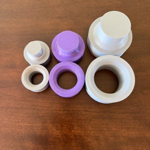 Może przedstawiać: Asortyment plastikowych przedmiot&oacute;w drukowanych w 3D o r&oacute;żnych kształtach i rozmiarach. Kolekcja obejmuje cylindryczne wtyczki i pierścienie w odcieniach szarości i fioletu. Obiekty są ułożone na drewnianej powierzchni.