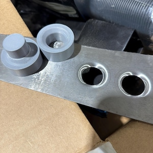 May include: Close-up of automotive parts. Two gray, cylindrical pieces sit next to a metal plate with two circular holes. The parts appear to be made of a 3D-printed material. A cardboard box is visible in the background.