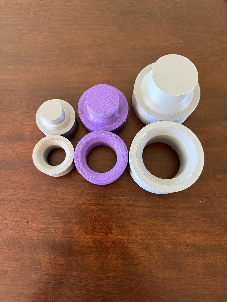K&ouml;nnte beinhalten: Eine Auswahl an 3D-gedruckten Kunststoffobjekten in verschiedenen Formen und Gr&ouml;&szlig;en. Die Objekte umfassen zylindrische Stecker und Ringe in Grau- und Lilat&ouml;nen. Die Artikel sind auf einer braunen Holzoberfl&auml;che angeordnet.