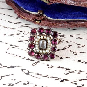 May include: A ring featuring a central rectangular stone surrounded by a border of small white seed pearls, enclosed within an outer square frame of deep red faceted garnet stones set in a metal band, showcasing a cluster design with intricate details.