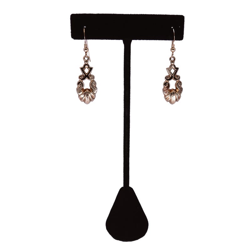May include: A pair of silver-toned earrings with a decorative design, displayed on a black jewelry stand. The earrings feature a floral-inspired shape with intricate detailing. The stand has a T-shape with a triangular base.