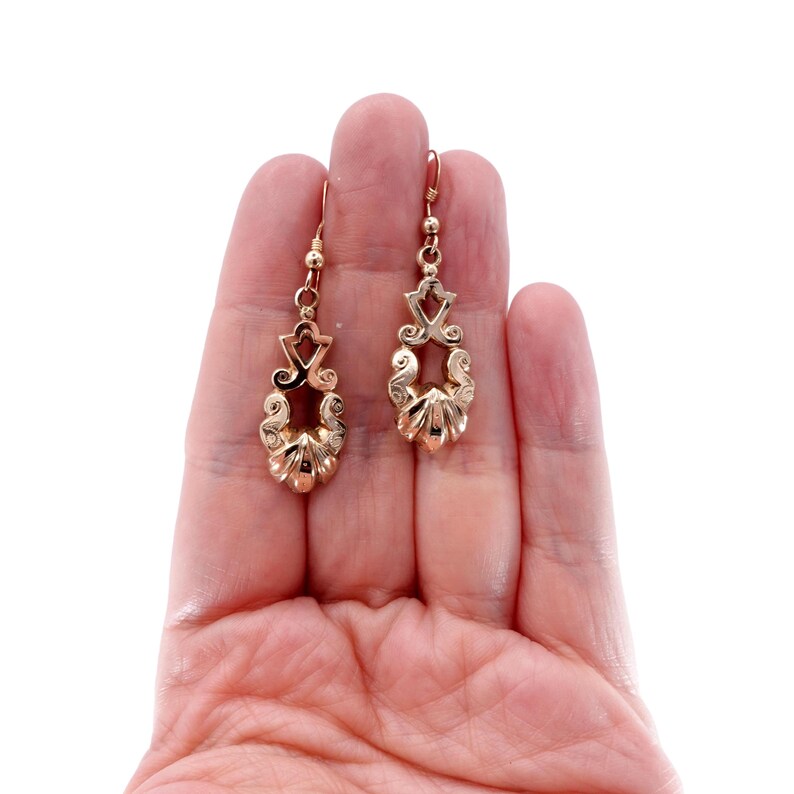 May include: A pair of gold-colored drop earrings. Each earring features an ornate design with a teardrop shape, intricate scrollwork, and a small hook for the ear. The earrings are held in a hand against a white background.