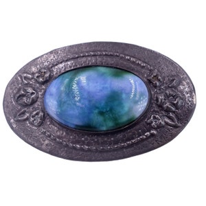 Pode incluir: Um broche oval com uma pedra de esmalte azul e verde engastada em uma moldura de metal prateado. A moldura é decorada com desenhos florais.
