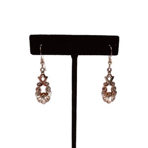 May include: A pair of silver-toned drop earrings. Each earring features a decorative design with a star-like shape at the top, a central openwork design, and a shell-like bottom. The earrings are displayed on a black stand against a white background.