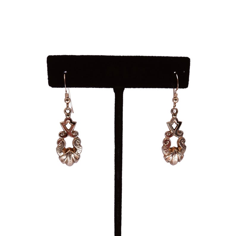 May include: A pair of silver-toned drop earrings. Each earring features a decorative design with a star-like shape at the top, a central openwork design, and a shell-like bottom. The earrings are displayed on a black stand against a white background.