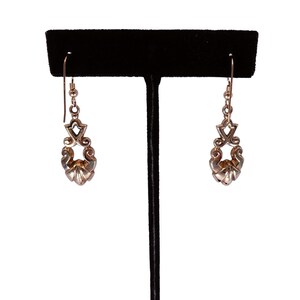 May include: A pair of silver-toned drop earrings. Each earring features an ornate design with a floral-like bottom and a decorative top section. The earrings are displayed on a black jewelry stand against a white background.