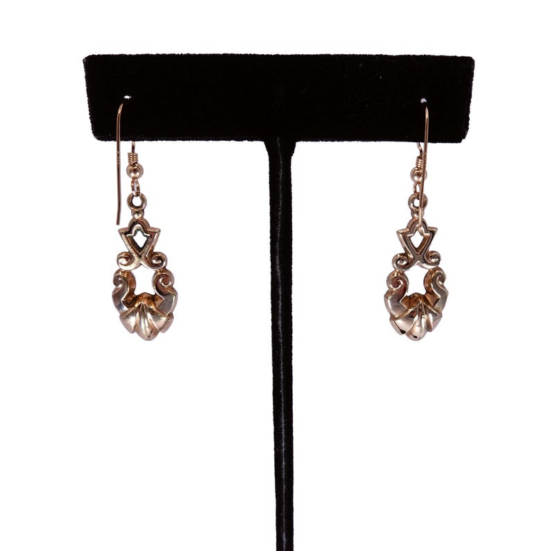 May include: A pair of silver-toned drop earrings. Each earring features an ornate design with a floral-like bottom and a decorative top section. The earrings are displayed on a black jewelry stand against a white background.