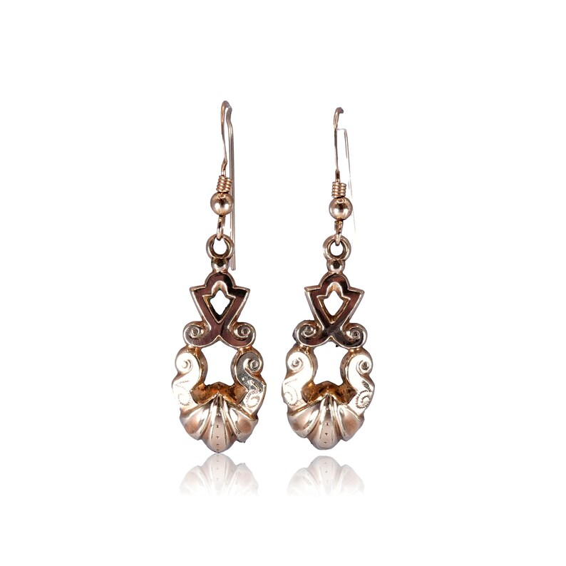 May include: A pair of silver-toned drop earrings. Each earring features an ornate design with a decorative top, a central openwork element, and a lower, shell-like shape. The earrings are suspended from a simple hook.