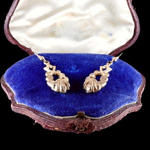 May include: A pair of gold-colored drop earrings displayed in an open vintage jewelry box. The earrings feature a decorative design with a teardrop shape and intricate detailing. The box has a blue velvet interior and a brown leather exterior.
