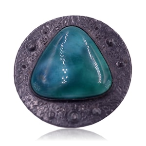 Pode incluir: Um broche prateado com um design floral intrincado que envolve uma gema verde triangular.