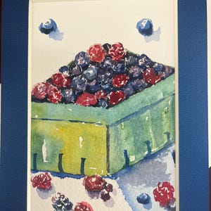 May include: Watercolor painting of a green berry basket overflowing with blueberries and raspberries. The artwork is framed in a white mat and a dark blue frame. Scattered berries are painted around the basket.