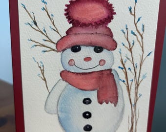 May include: Watercolour painting of a snowman wearing a pink hat and scarf, with black button eyes and a carrot nose. The snowman is on a white background with brown branches and blue accents. The card has a burgundy border.