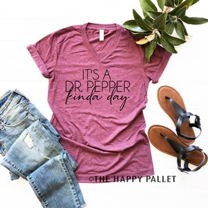 Puede incluir: Una camiseta de cuello en V color burdeos con el texto "It's a Dr. Pepper kinda day" impreso en negro.